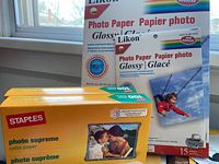 Photo of Staples Photo Supreme satin paper box with 100 sheets and Likon Photo Paper Glossy packages against window background.