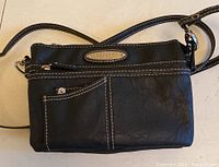 Black Rosetti crossbody purse with front zipper pocket and external stitching laid flat on a table.