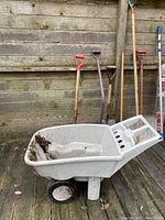 Grey plastic wheelbarrow with moss and dirt on the inside, resting on a wooden deck in front of a wooden plank wall. Several garden and snow tools lean against the wall nearby.