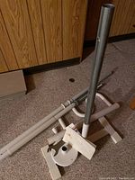 Photo of metal poles and mounting plate parts on carpeted floor with a wood-paneled wall background. Shows poles with white ends and mounting hardware.