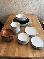Various stacked bowls and plates on a wooden table including white and black ceramic pieces and wooden bowls.
