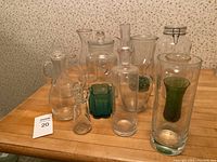 Clear and green glass vessels including pitchers, vases, jugs, and a large glass jar with clip lid on wooden table.