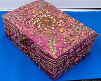 Closed purple fabric jewelry box with detailed gold and multicolor embroidery and beading, displayed on a blue surface.