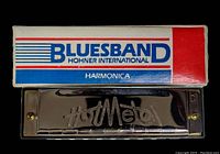 Photo of the harmonica with the shiny metal casing labeled 'Hot Metal' alongside its original box with 'BLUESBAND HOHNER INTERNATIONAL HARMONICA' branding.