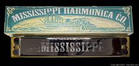 Top view showing vintage Mississippi Harmonica Co blue-green box with steamboat illustration and the harmonica in front with 'MISSISSIPPI' printed in white on black body.