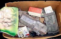 Box containing several gray and red model railroad building parts, white building, black tracks, and a green landscape piece.