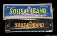 Top view of the black harmonica with gold 'Sousa Band' logo on the cover and original blue and white box with brand name and model number