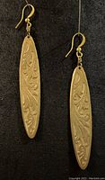 Pair of antique brass earrings with floral pattern detail on elongated oval shape, hanging on gold plated ear wires against black fabric background.