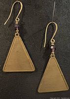 Close up front view of one triangular brass earring showing ribbed linear texture and purple crystal bead on gold plated hook.