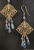 Pair of antique brass earrings with intricate diamond shaped textured pendants and frosted blue glass beads on black background.
