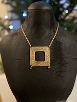 Front view of the antique brass necklace displayed on a black mannequin, showing the square pendant with drilled symbols and glass bead accents.