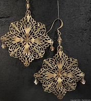Pair of large antique brass filigree snowflake earrings hanging against a black background, showing the overall size and design of the earrings including dangling crystal beads.