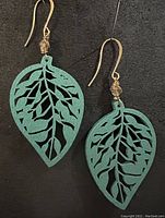 Pair of turquoise painted wood leaf earrings with intricate cutout leaf patterns on black background, showing overall design and materials.