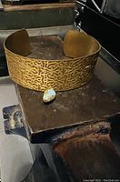 Full view of the antique brass cuff with patterned design and blue glass bead on a rustic surface.