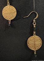 Close-up image of one earring showing the gold plated brass coin pendant with intricate scrollwork pattern and small dark blue Swarovski crystal at the bottom, wire wrapped detail, hanging from gold plated brass hook.