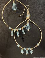 Full view of both gold plated brass hoop earrings with wire wrapped blue glass and crystal accents suspended on gold plated ear wires.