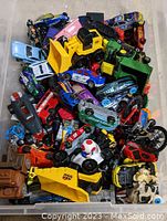 Top view of a clear plastic bin filled with assorted diecast toy vehicles including cars, trucks, motorcycles, and construction equipment in various colors.