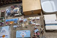 Overview photo showing all items in the lot arranged on floor including sewing supplies, wooden box, strainer, sheet set, vacuum bags, figurines and packaged birds.