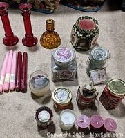 Photo shows assorted candles including tapered candles, jar candles with lids, smaller votive and pillar candles, three candlestick holders (two red glass, one amber glass), and a ceramic candle warmer with green floral pattern and red insert.