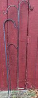 Photo of three black metal shepherd's hooks against a red wooden background, including a 7-foot double hook, a 6-foot single hook, and a 5-foot single hook.