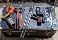 Photo showing the open trunk with various garden tools arranged on top: plunger, bulb planter, hand rake, pruning shears, hose nozzle, hammer, trowel, and ice scraper in packaging.