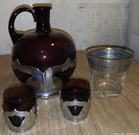 Photo showing a dark-glazed ceramic decanter in a metal holder, two matching small glasses with metal holders and a decorated clear glass with silver pattern rim.