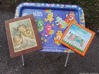 Photo shows a Care Bears themed metal TV tray with foldable legs standing against a concrete wall, leaning on grass. Next to it is a framed print of two children and a zippered wall pocket organizer with vegetable garden print.