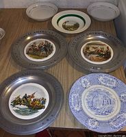 Photo showing seven collector plates including four plates commemorating the Great American Revolution 1776 featuring detailed historical scenes in color, two plates with blue and white floral or building patterns, and one plate with a green rim depicting an animal illustration.
