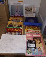 Photo of crafting books, iron-on transfers, and large craft patterns stacked on the floor, showing colorful book covers and transfer packages.