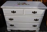 Front view of white dresser showing four drawers with floral decal on top surface and metal handles.