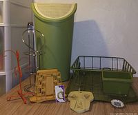 Photo showing a variety of kitchen items including a green basket with an attached small container, a tall green container with perforated lid, an orange utensil holder, a rotating metal stand, a wooden slicer, a small roll of tape, a beige organizer, and a floral trinket dish