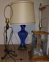 Photo showing all three lamps together on a surface, including a blue glass lamp with beige shade, wood and glass lamp with nature scene, and black metal sculptural base lamp.