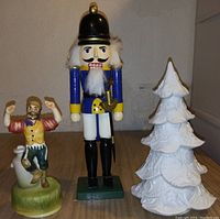 Photo showing three figurines: a wooden nutcracker soldier, a ceramic music box figurine of a man with raised hands, and a white ceramic tree decoration.