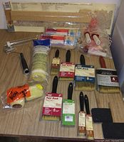 Full view of lot showing paint brushes of various sizes, roller covers, and applicators on wood table surface