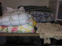 Folded bedding items arranged on a surface including floral sheets, multiple blankets in different colors and patterns, and a crochet lace piece.