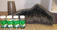 Photo showing a gray plush triangular small pet bed, four green catnip bottles labeled Catnip N Wonders Catnip Mix, and a brown plastic flower pot on a wooden surface.