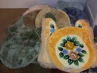 Photo shows five toilet seat covers and bath rugs in various shapes, colors and a floral design on one cover.