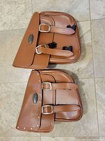 Two brown leather motorcycle saddlebags with buckle straps and black clasps shown on tiled floor.