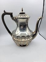 Side view of antique silver coffee urn showing wooden handle, ornate body details, curved spout, and decorative lid with finial