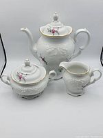 Front view of Royal Kent porcelain teapot, creamer, and sugar bowl set with floral embossing and gold trim.