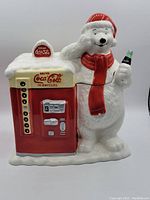 Front and side view of the vintage Coca-Cola polar bear cookie jar holding a soda bottle and standing next to a vending machine replica.