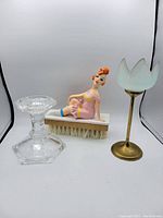 Overall view of the three items in the lot: clear glass candle holder, vintage figurine brush with girl in pink dress, and brass tulip votive candle holder with frosted glass top.