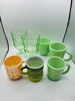 Front view of all 8 items together: 3 Fire-King jadeite mugs, 2 clear green glasses, 1 orange textured mug, 1 green textured mug.