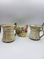 Two vintage speckled beige pottery mugs with flared rims and vertical panel design flanking a ceramic chicken coin bank with chicks