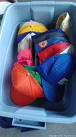 Photo showing multiple baseball style hats inside a blue plastic bin from above, displaying various colors and styles.