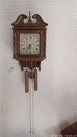 Full front view of wall clock hanging on wall showing wood case, dial, weights and pendulum