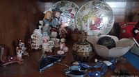 Overall view of lot with figurines, plates, metal apple, bowl, bell, and vase arranged on wood shelf