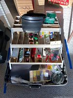 Open fishing tackle box showing multi-tiered trays with fishing thread spools, feathers, fly-tying supplies, magnifier headpiece placed on top, and various fishing accessories.