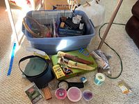 Wide view of fishing supplies arranged around and inside a large blue plastic storage bin on carpeted floor, showing knives, storage cases, fishing line, hooks, and other fishing accessories.