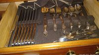 Drawer of flatware showing knives, forks, small decorative forks, and spoons arranged in fabric slots inside wooden drawer